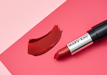 A red Mary Kay lipstick is photographed with its cap off laying atop a pink background and alongside a smear of lip color.