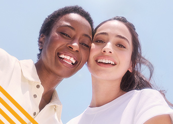 Two women with diverse skin tones, one with dark skin tone, the other with medium skin tone, are standing against a bright blue background. Both women are smiling and radiating positivity.