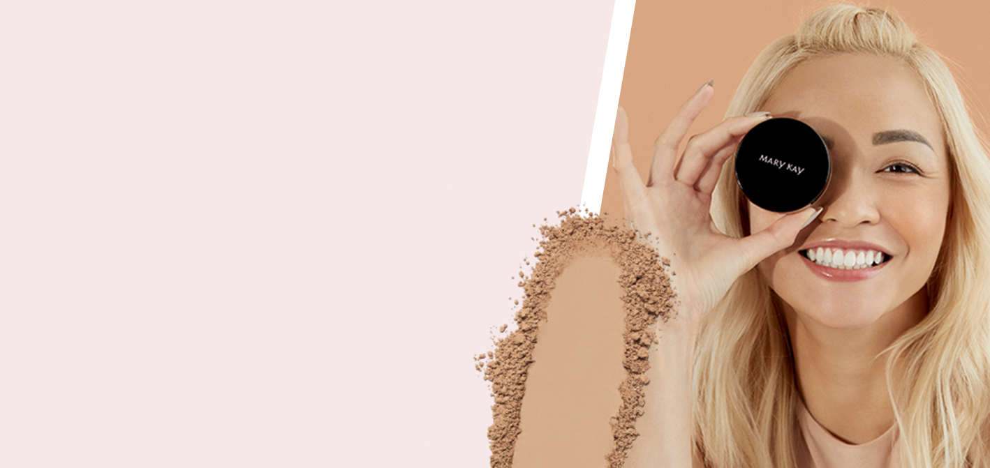 A blonde, Asian Mary Kay model in front of a half pink, half tan background and behind a smear of powder holds a jar of Mary Kay Silky Setting Powder over one eye and smiles.