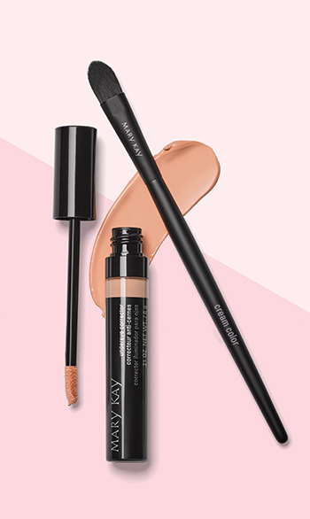 An open tube of Mary Kay Undereye Corrector is photographed alongside its doe-foot applicator and a cream color makeup brush and atop a product smear in front of a two-toned pink background.