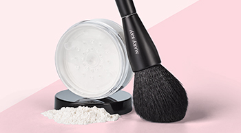 An open jar of Mary Kay Translucent Loose Powder is photographed with powder spilling out alongside an all-over powder makeup brush in front of a two-toned pink background.