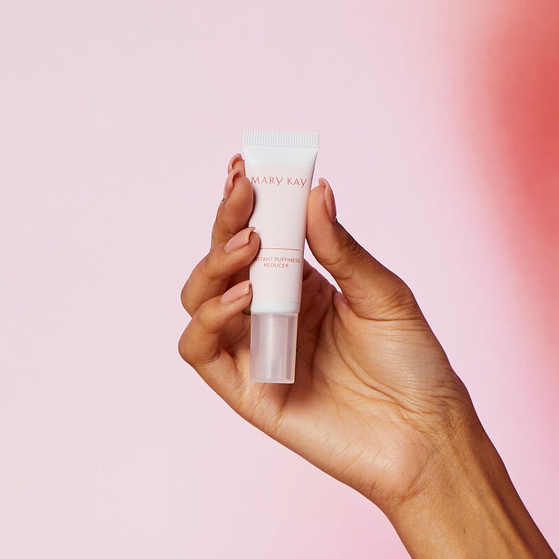 Hand holding Mary Kay Instant Puffiness Reducer against pink gradient background