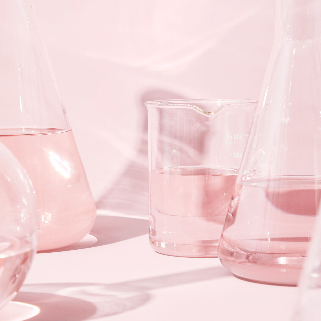 Des b&eacute;chers transparents remplis de liquide incolore sont pr&eacute;sent&eacute;s sur une surface rose qui refl&egrave;te un &eacute;clairage rose p&acirc;le.
