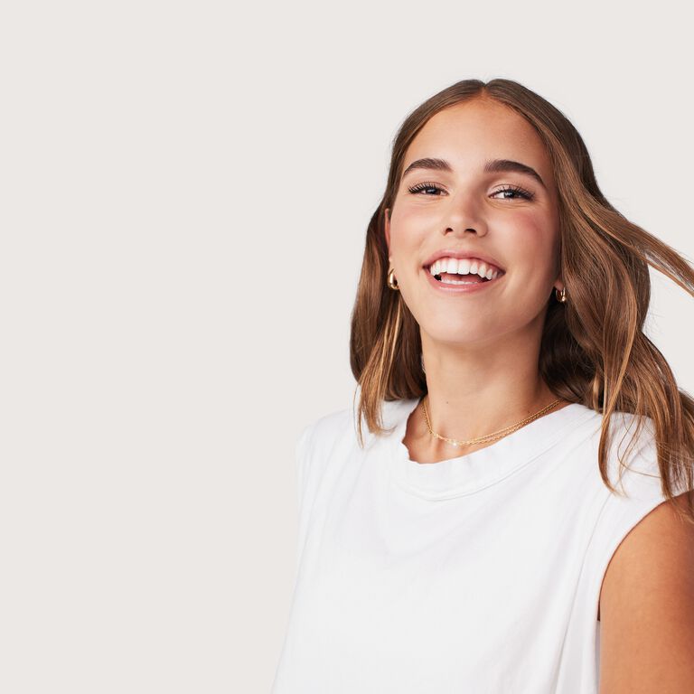 Smiling person with brown hair, wearing a white top and gold necklace, against a plain light background