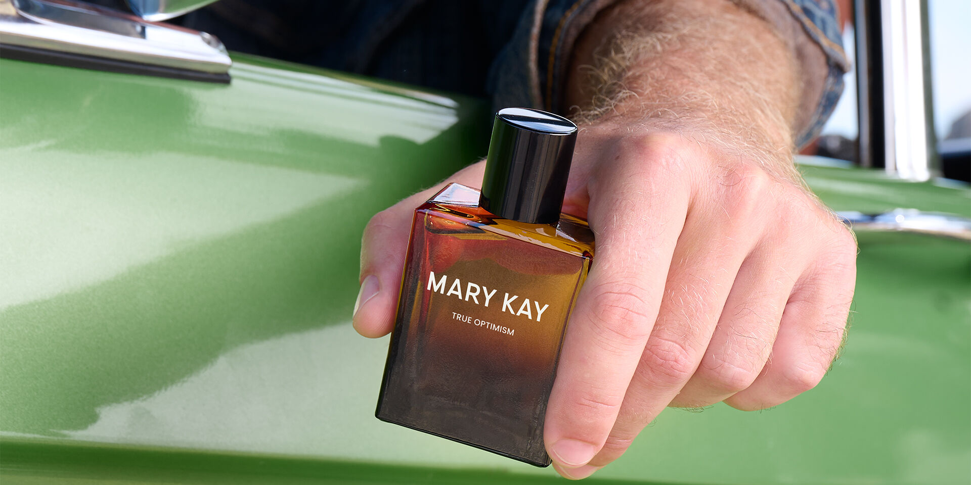 La main d&rsquo;un mannequin &agrave; la peau claire tient le parfum Mary Kay&reg; True Optimism&trade; Eau de Parfum, visible &agrave; travers la vitre d&rsquo;une voiture verte